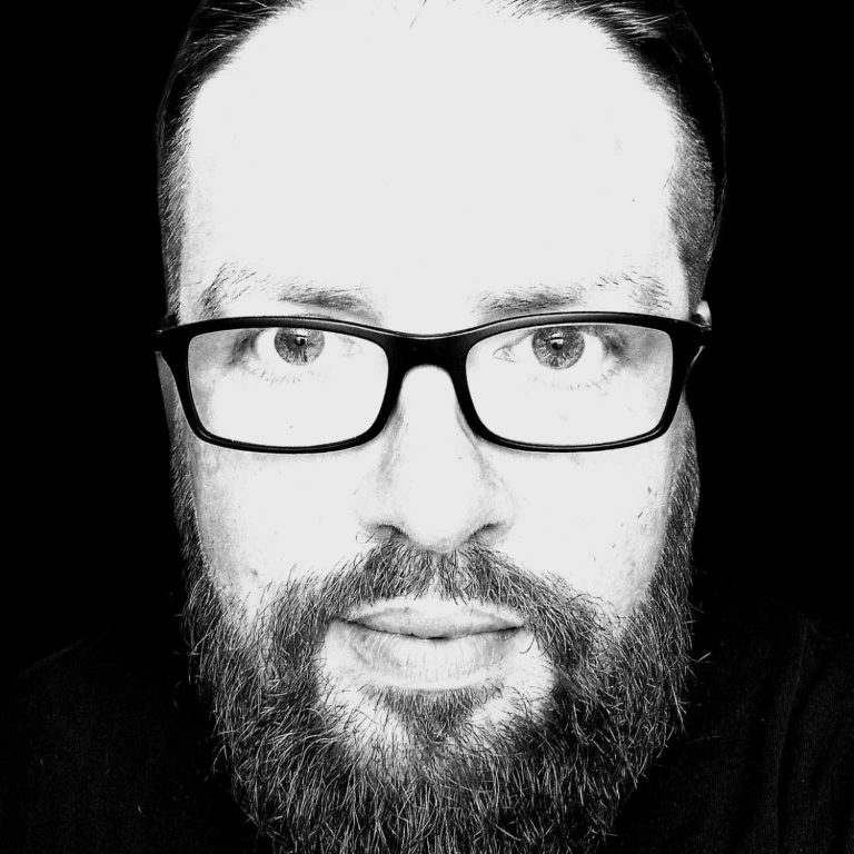 Foto von DJ MicDi Mann mit Brille und Vollbart vor schwarzem Hintergrund, direkt in die Kamera schauend.