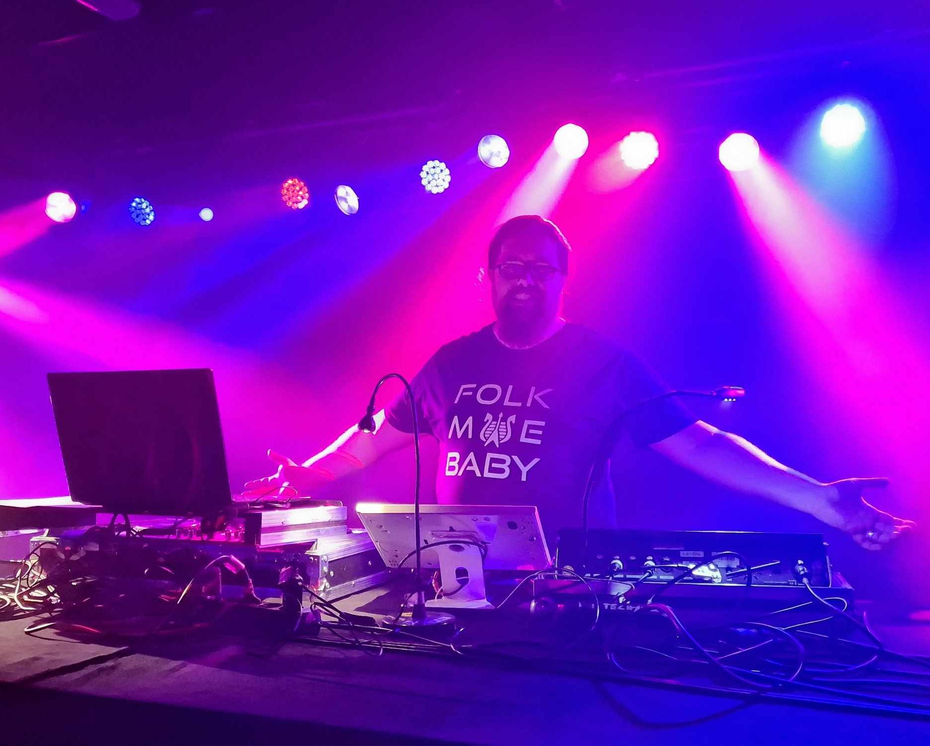 DJ MicDi steht an einem Mischpult DJ MicDi steht an einem Mischpult mit bunten Lichtern und hebt die Hände.